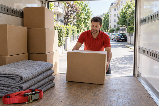 Un déménageur en polo rouge soulève un carton au hayon du camion, des couvertures pliées à ses pieds.