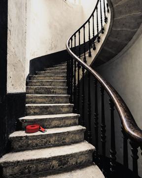 Une cage d’escalier en colimaçon, rampe de fer forgé, une sangle rouge posée sur une marche.
